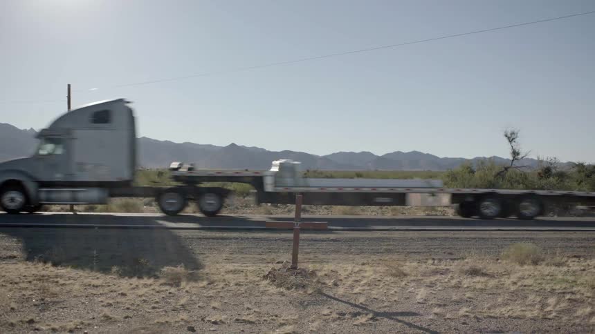 A semi-truck drives by a wood cross next to a road in the desert