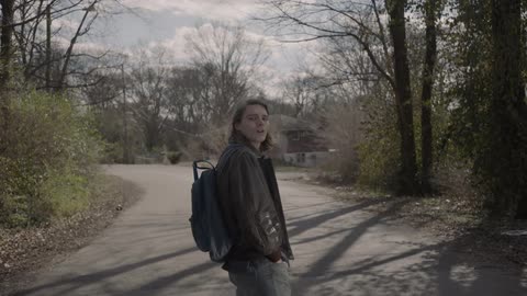 A man with a coat and backpack on is waiting on a road staring back at the camera