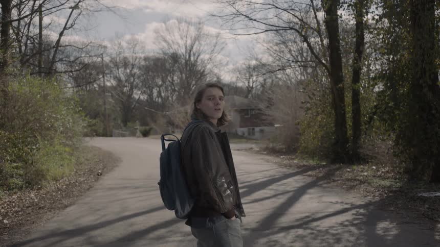 A man with a coat and backpack on is waiting on a road staring back at the camera