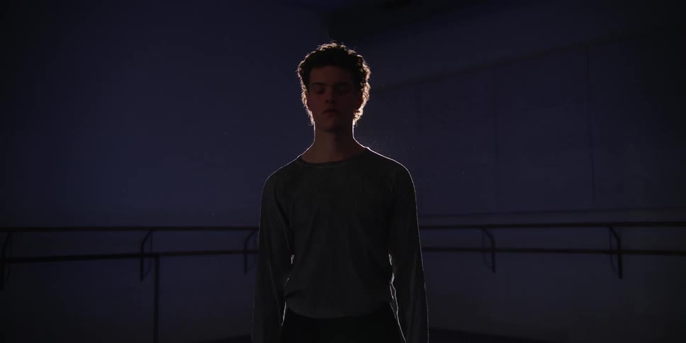 Male dancer stretches while back lit by spotlight in dim dance studio