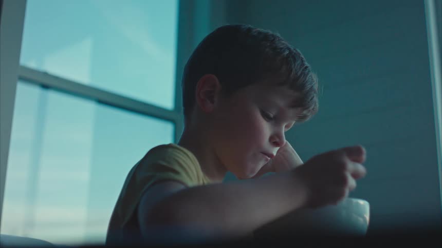 Boy sits and eats while thinking at table near window