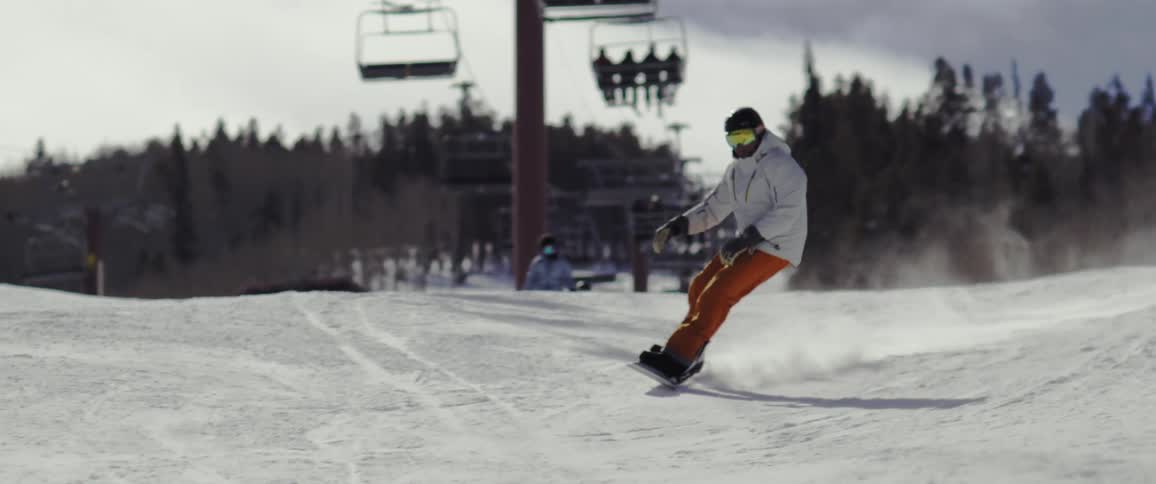 A person in white clothes is skiing on the snow under a moving chairlift and behind is a pine forest