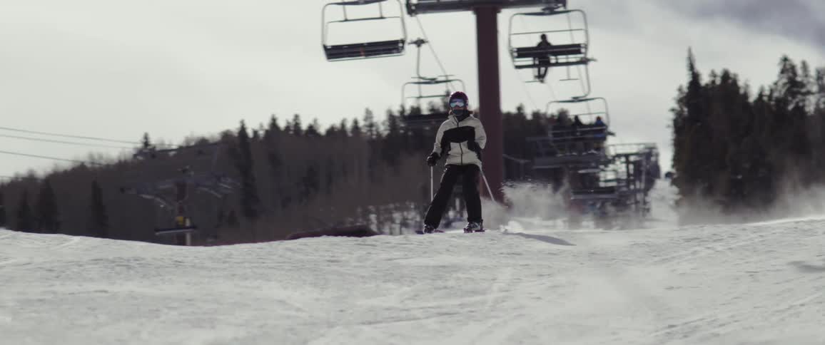 A person skiing on the snow under a moving chairlift and behind is a pine forest
