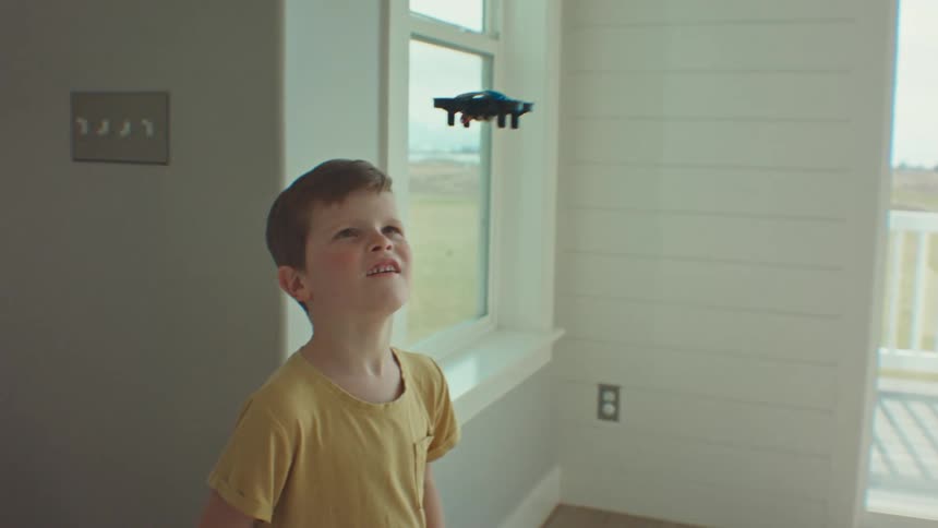 A boy wearing a yellow shirt flies a drone indoors above a table near windows