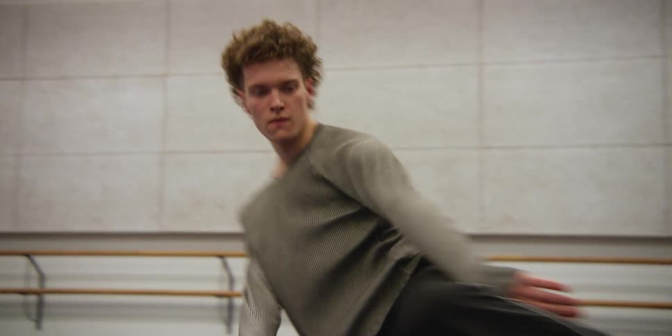 A curly-haired young adult male dancer practices spinning on a dance floor.