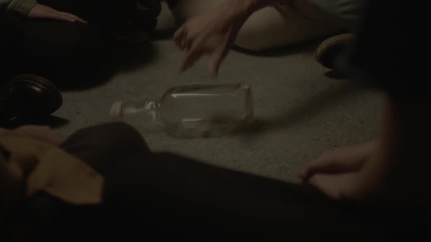 A group of people surround a glass bottle as it is spun on the floor