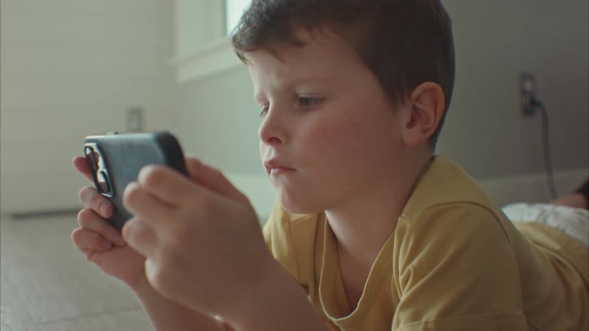 A little boy is lying down and is playing with a cell phone with his hands and is looking intently at it