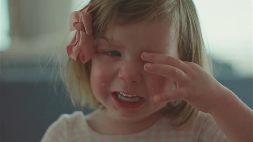 A crying toddler rubbing her eyes while standing indoor at midday