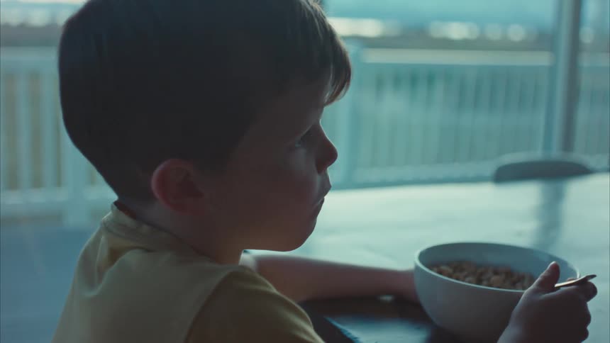 A little boy with his back turned is eating cereal from a bowl using a spoon