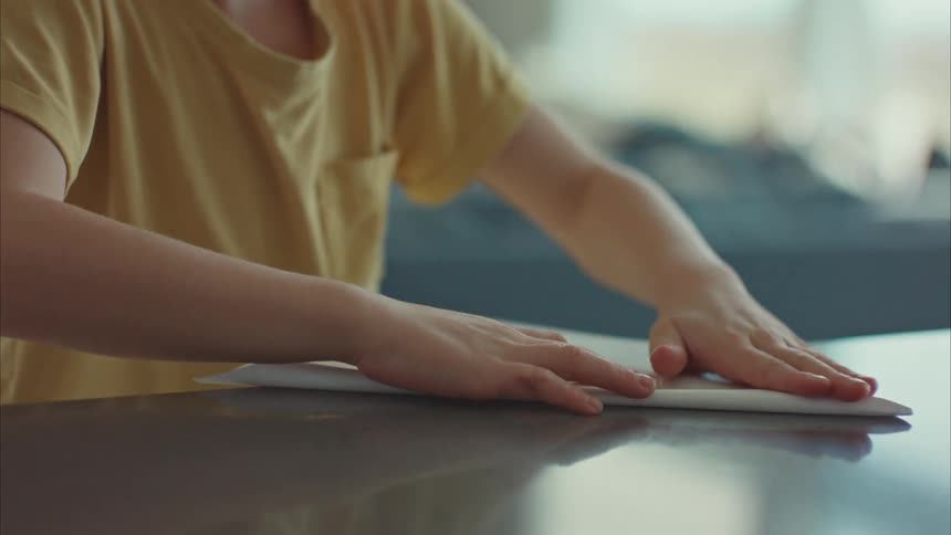 A little boy is assembling a paper airplane on a table that is reflecting the light of the room