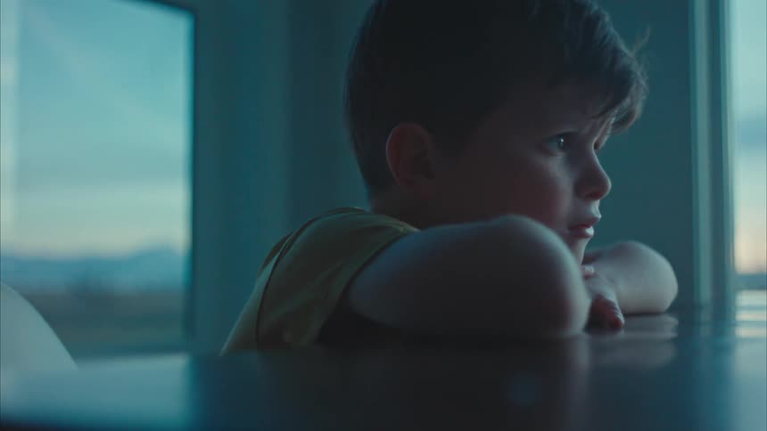 A little boy has his head resting on his arms which are on a table
