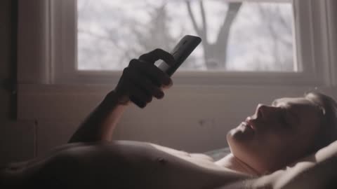 A man without a shirt is laying in bed checking his phone