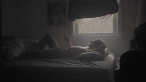 Shirtless man lays on bed looking at his phone