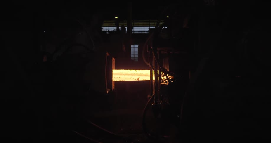 A glowing hot metal beam pushing from one machine into another in a dark steel factory