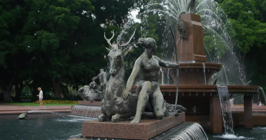 Pedestrians move through park near large water fountain containing multiple statues