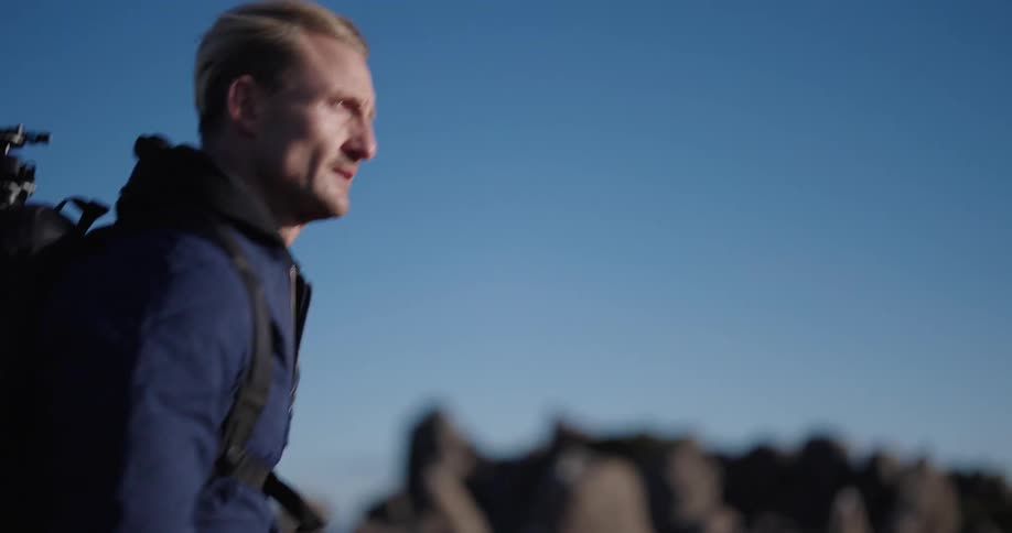 Male photographer hikes past boulders carrying backpack containing a tripod