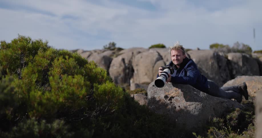 A man is laying on a rock with a camera in hand on top of a mountain