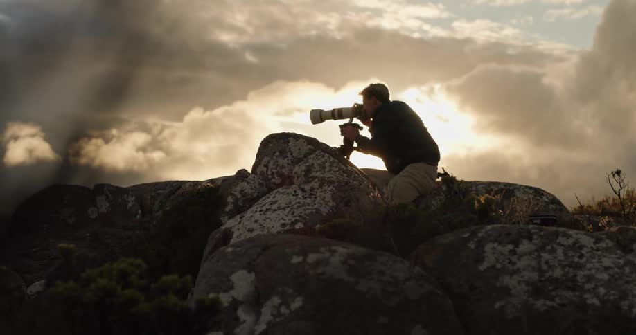 Male photographer sits amongst boulders taking picture with a telephoto lens on a cloudy day