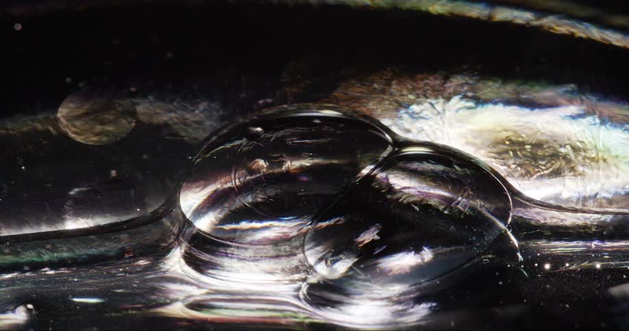 Extreme closeup of one bubble joining another amidst a shiny liquid