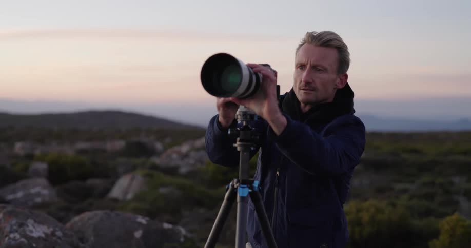 A man outdoors with a camera on a tripod capturing the landscape during dusk