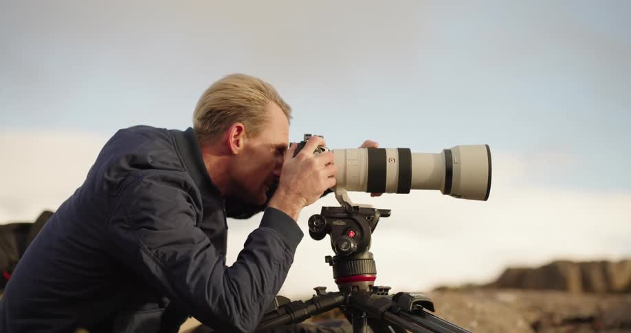 A blond man is focused with his face on a camera that is on a tripod and the camera has the lens zooming in