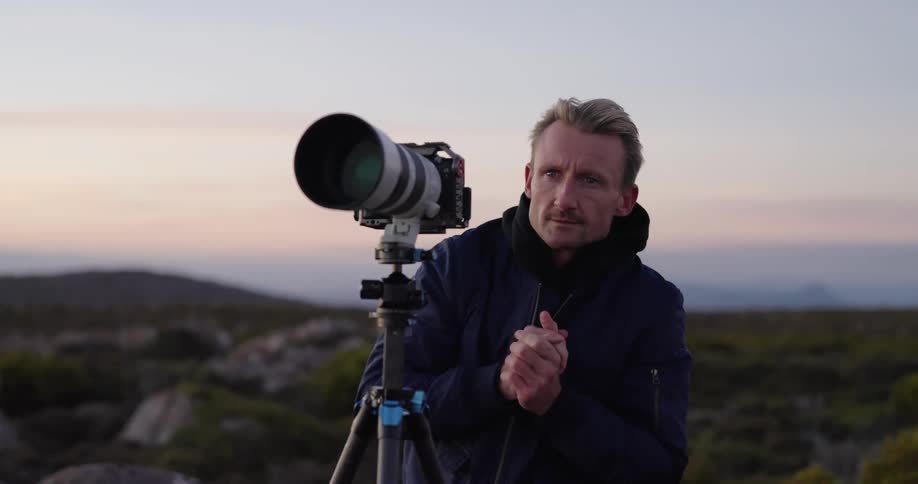 Photographer stands behind telephoto lens on tripod outdoors at dusk