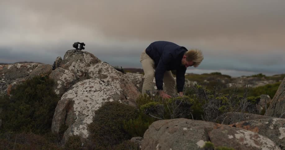 A photographer outdoors stores a camera in his backpack on a rocky landscape