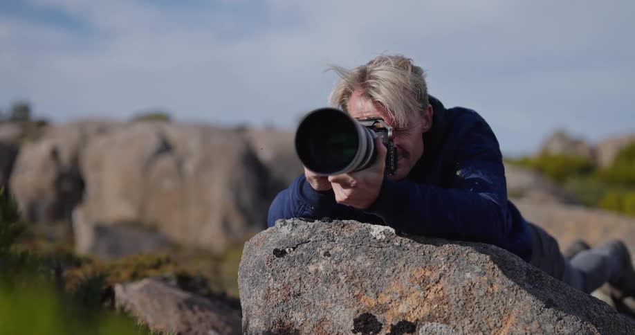 Male photographer lying against boulder takes photograph using a camera with a telephoto lens