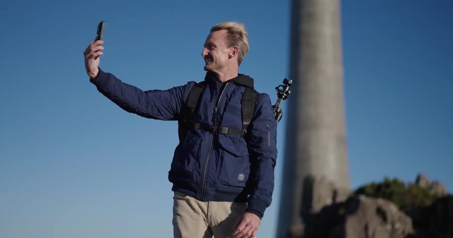 Men wearing blue jacket and backpack takes selfie outdoors under blue sky