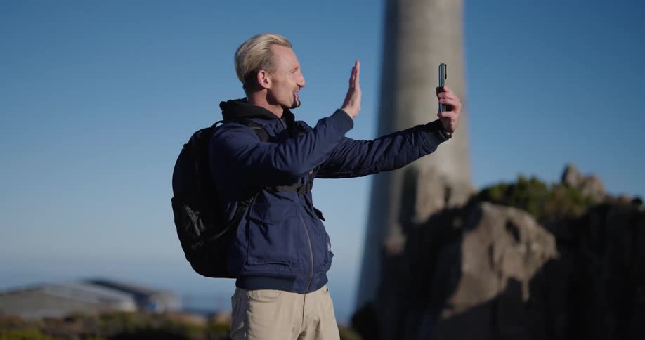 Man wearing jacket and backpack takes selfie outdoors and waves at camera