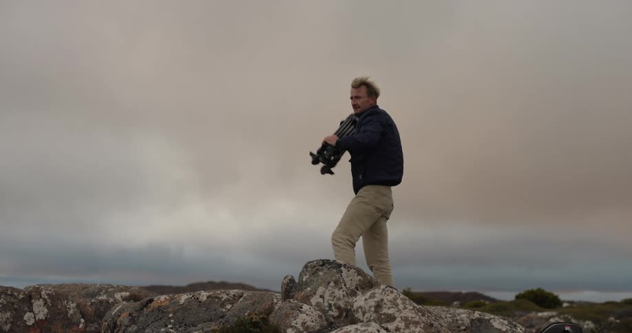 A man picks up a tripod from the ground and puts it on his back and carries it walking to the right in a region where there is a lot of wind that is messing with his clothes