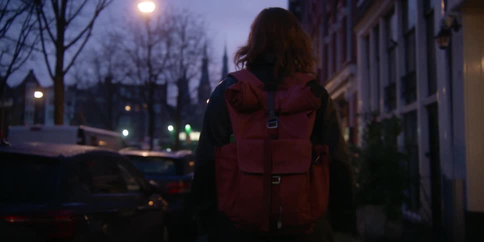 A woman with a backpack glances to the side at lit buildings and vehicle traffic as she walks down a city sidewalk at dusk.