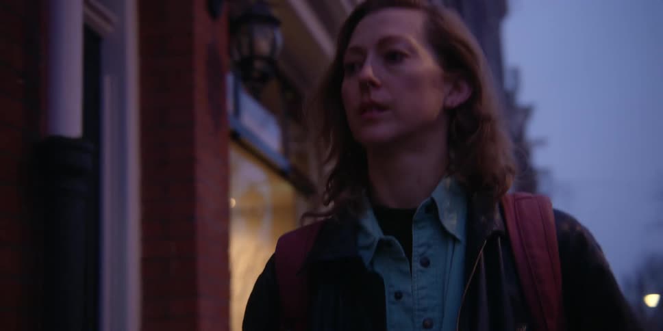 A fair, red-headed woman wearing a backpack walks down the sidewalk in the early morning hours, then turns to the door of a brick, windowed building.