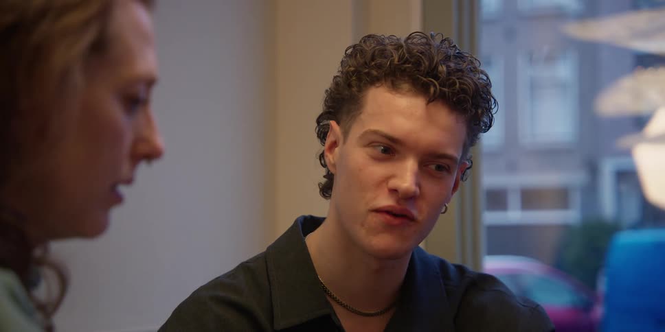 A curly-haired young male client and a red-headed adult woman, both dressed alternatively, sit together at a table viewing a laptop and discussing a project.