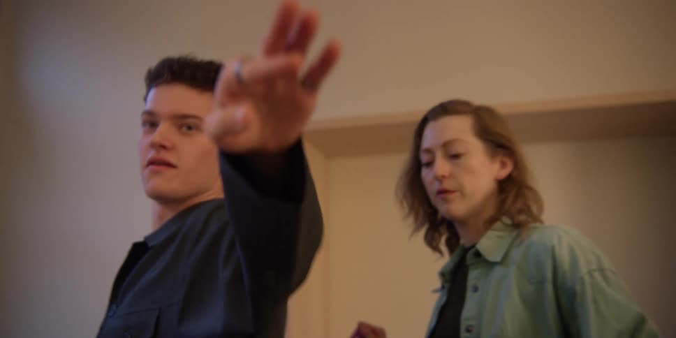 A red-headed female dance instructor works with a young, curly-haired Caucasian male on his arm, hand, and body movements.