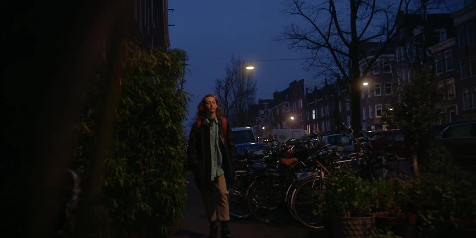 An adult woman passes a child going to school as they both walk on a city sidewalk full of bikes and parked cars in the early morning hours.