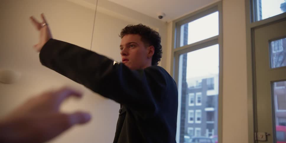 A young male dancer practicing his dance techniques in a city apartment studio as an off-screen female instructor demonstrates hand and arm movements.