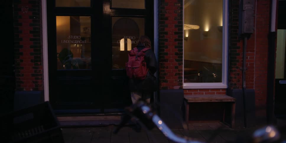 POV from behind bike handlebars of a woman with a backpack walking on a city sidewalk before dawn to a windowed small business where she unlocks the door and lets herself in.