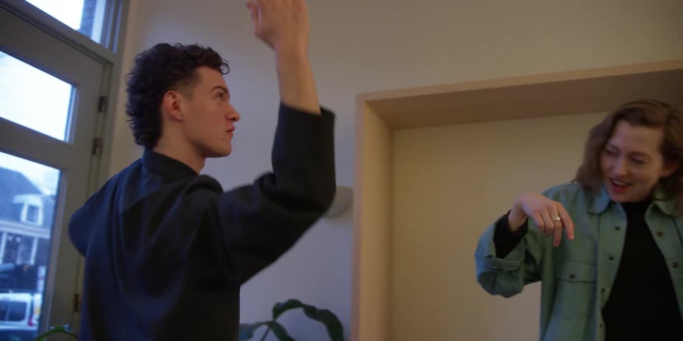 A curly and dark-haired white male dancer practices his arm and body movements as he is coached by an older, white female dance instructor.