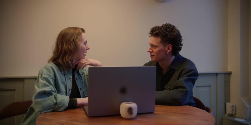 A red-headed adult woman and a young curly--haired adult male view something on a laptop and discuss what they are seeing.