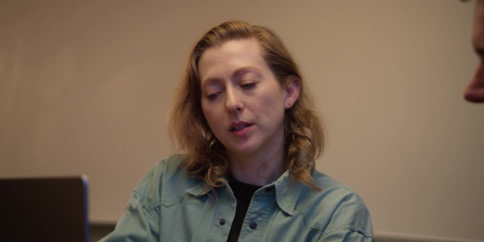 A middle-aged woman with fair skin and light eyes sits next to a young adult male with curly hair as they interact expressively while viewing something on a laptop.