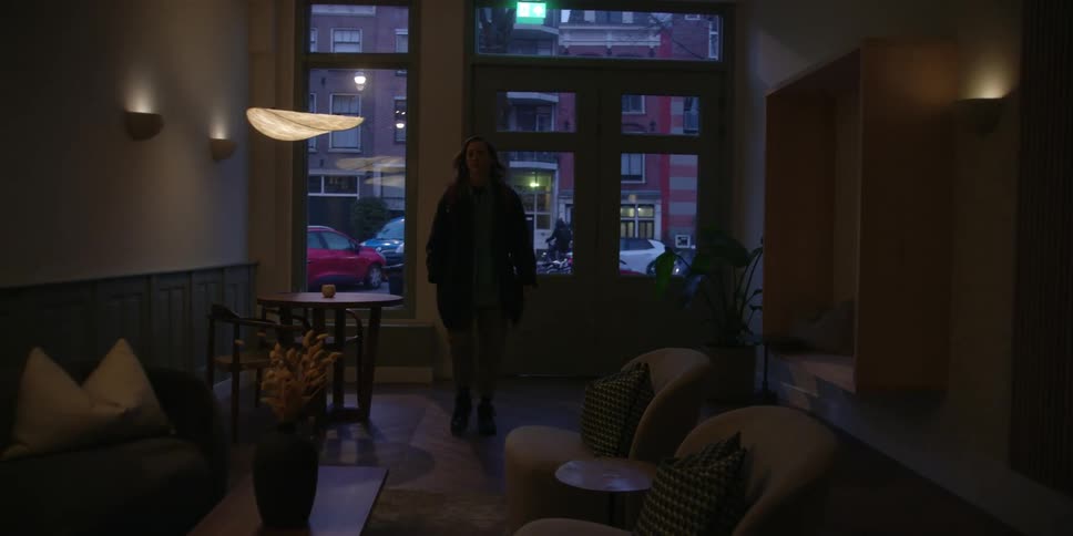 A woman walks in the front door of a studio with windows to the city outside and walks through the seating area to turn up the lights.