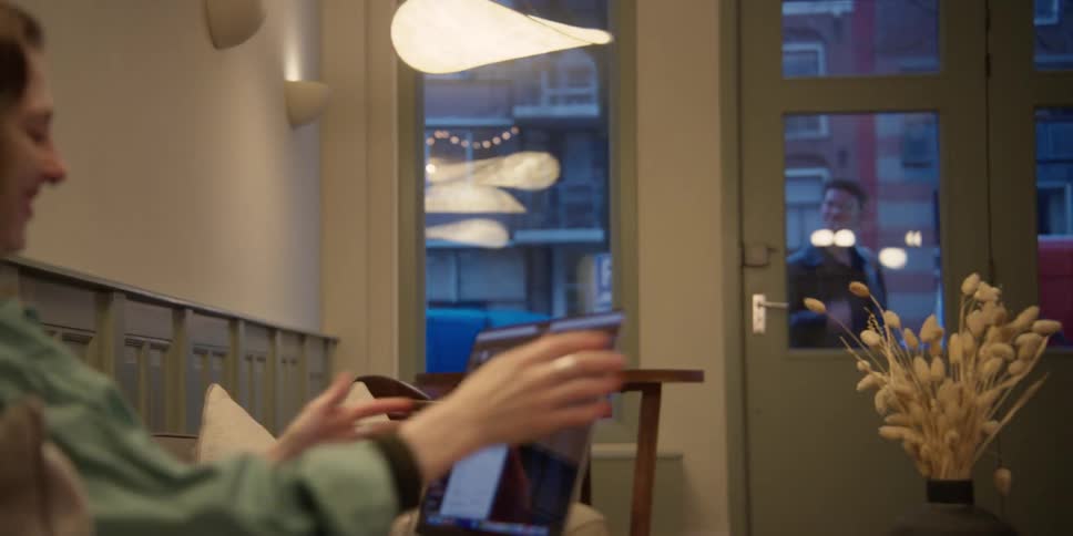 A woman with red hair and septum ring is engrossed in her laptop when she notices and waves to her young adult male friend outside the window and gets up to let him in the glass studio door.