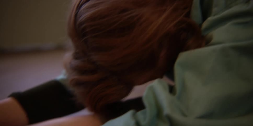 An adult woman with red-hair and a nose ring is sitting and stretching her neck, arms, legs, and body.