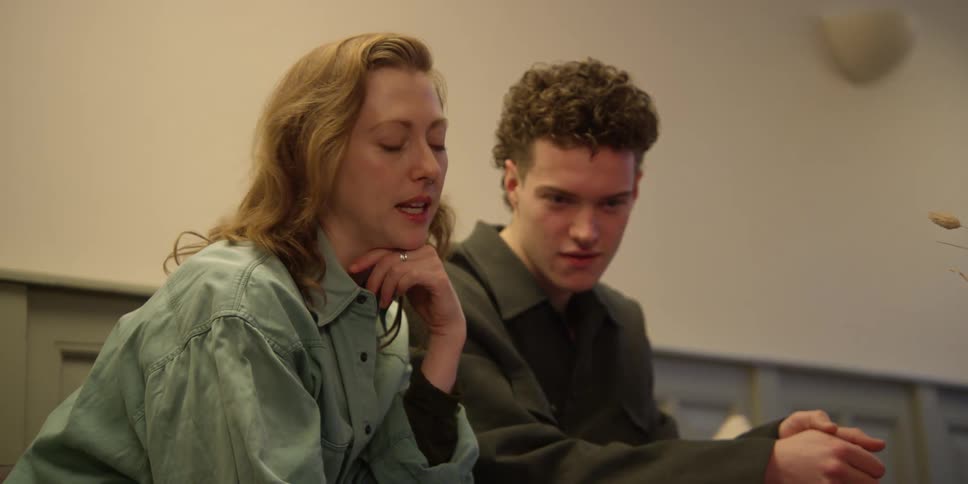 A red-headed woman with a septum ring leans on her hand and fiddles with her laptop off screen while talking and gesturing with the young curly-haired man sitting beside her as they discuss what they are seeing.