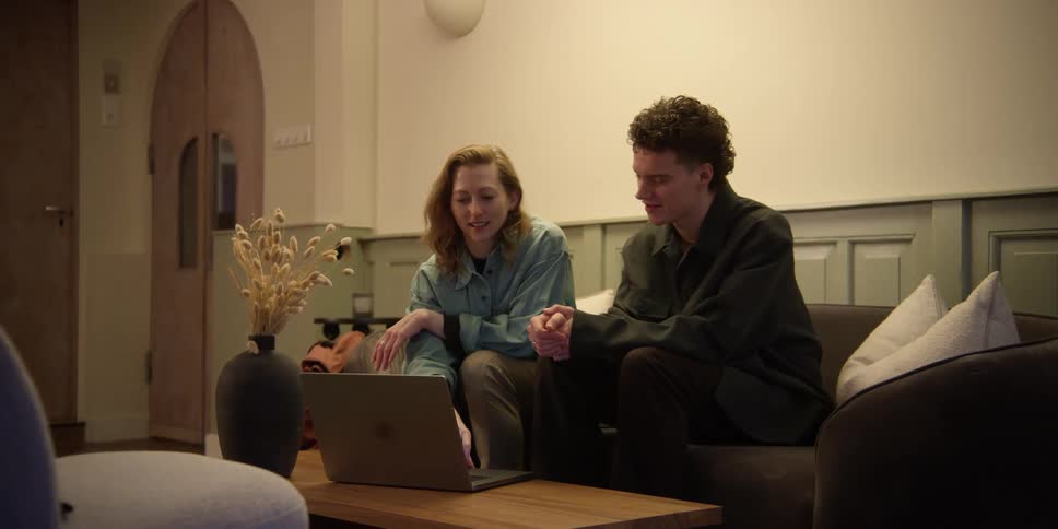 A young curly-haired man and adult ginger-haired woman are sitting on a couch and looking at a laptop while talking and gesturing about what they're viewing.