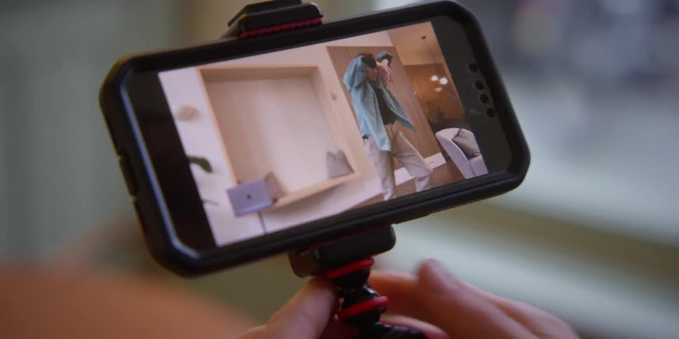 A person holding their phone and watching a video of a woman practicing a dance in her living room.