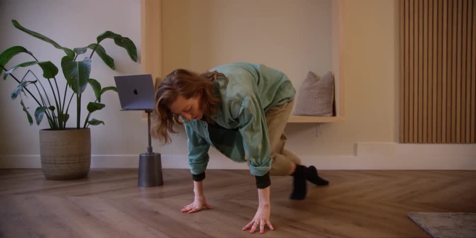 A fair red-headed woman is doing leg and arm stretches indoors on a hardwood floor with a laptop on a stand behind her