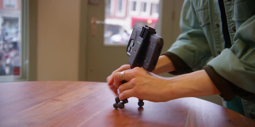 A woman in an apartment with windows to the city outside sets up a smartphone on an adjustable mini tripod to record.