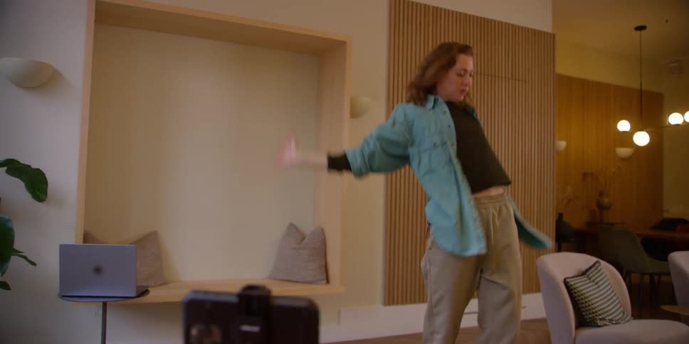 A female dancer practices contemporary dance movements in her living room while she films herself with her smartphone on a stand.